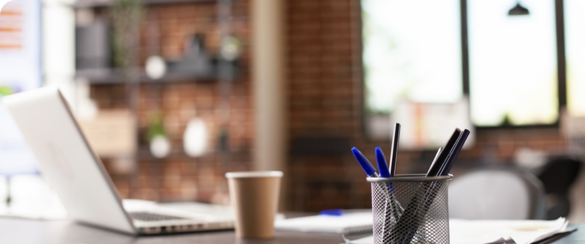 Laptop, Kaffeebecher und Stifte auf Schreibtisch im Büro