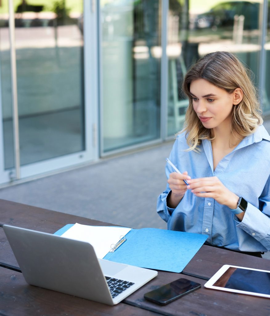 Frau arbeitet am Laptop im Außenbereich, mit Ordner und Tablet auf dem Tisch.